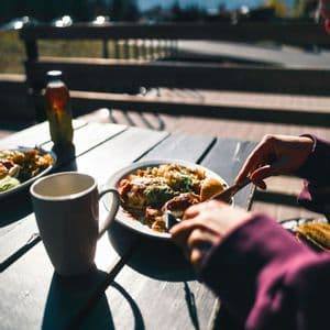 Una persona mangia un pasto da un piatto bianco a un tavolo all'aperto, con una tazza e montagne sullo sfondo.
