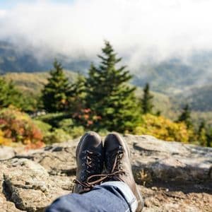 Le gambe di una persona con scarponi da trekking riposano su una sporgenza rocciosa, con vista su un paesaggio montuoso con alberi sempreverdi e nuvole basse.