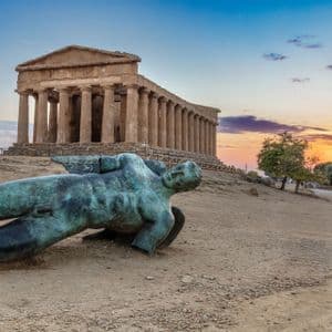 A bronze statue of a fallen winged figure lies on a dusty hill with an ancient Greek temple in the background at sunset.
