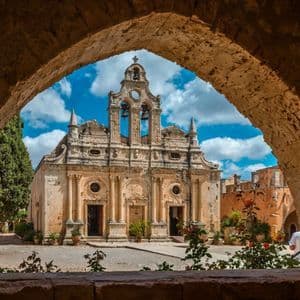 An ornate stone monastery with a bell tower sits in a courtyard, viewed through a weathered stone arch under a partly cloudy sky.