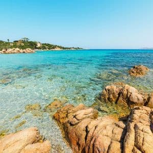 Acqua limpida e turchese lambisce una costa rocciosa con vegetazione verde sotto un cielo azzurro brillante.