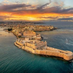 An aerial view of a historic coastal city and a stone fortress on a peninsula under a dramatic sunset sky.