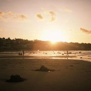 Sagome di persone su una spiaggia sabbiosa e nell'oceano durante un tramonto dorato su una costa collinare.