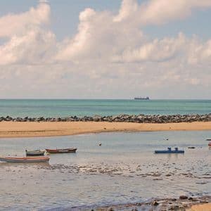 Piccole imbarcazioni sono ormeggiate in una tranquilla insenatura, di fronte a una spiaggia sabbiosa e a una barriera frangiflutti in roccia, con una nave da carico all'orizzonte.