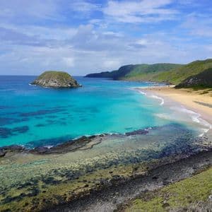 Una vista panoramica di una baia tropicale con acqua turchese, una spiaggia sabbiosa e colline verdi sotto un cielo azzurro con nuvole.