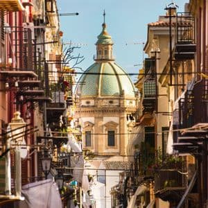 A green-tiled church dome is framed by tall buildings on a narrow street with laundry hanging from balconies.