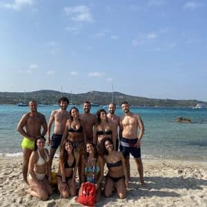 A WeRoad group trip in swimwear posing on a sandy beach in front of turquoise water with boats.