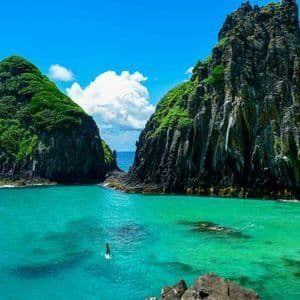 Una persona in stand-up paddleboard scivola su acque turchesi tra due grandi formazioni rocciose ricoperte di verde.