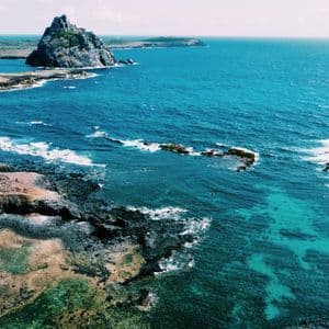Vista aerea di una costa frastagliata con un'isola rocciosa che si staglia in un vivace oceano turchese.