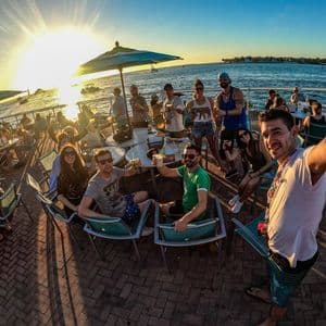Un grupo de WeRoad se toma una selfie en una terraza junto al mar al atardecer, con bebidas en sus mesas.