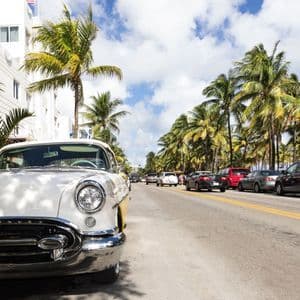 Un coche antiguo blanco aparcado en una calle soleada bordeada de altas palmeras junto a un edificio blanco.