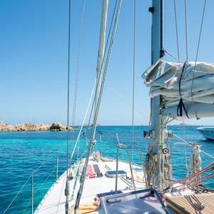 Vista dal ponte di una barca a vela su acqua cristallina turchese verso una costa rocciosa sotto un cielo azzurro brillante.