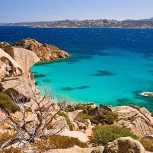 Una vista panoramica da una costa rocciosa che si affaccia su una baia con acqua cristallina color turchese, con il mare blu intenso e una costa lontana sullo sfondo.