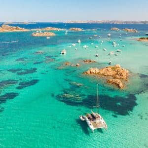 Vista aerea di un catamarano e altre barche ancorate in una baia turchese poco profonda tra isole rocciose.