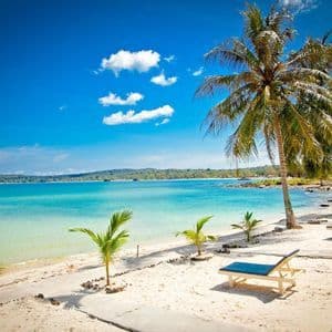 Ein sonniger Tropenstrand mit weißem Sand, Palmen und Liegestühlen an ruhigem türkisfarbenem Wasser.