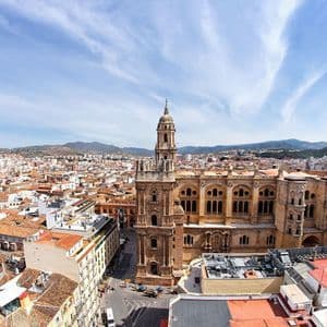 Une vue aérienne d'une cathédrale historique entourée d'un paysage urbain avec des toits de tuiles rouges et des montagnes en arrière-plan.