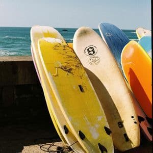 Una fila di tavole da surf colorate è appoggiata a un muretto di cemento, con l'oceano blu visibile sullo sfondo.