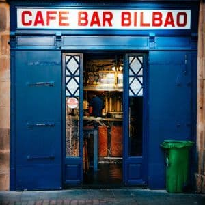 La vetrina blu del Cafe Bar Bilbao, con un'insegna illuminata e le porte aperte su una strada di ciottoli.