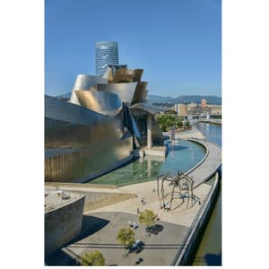 Una vista aerea di un moderno edificio metallico e una grande scultura di ragno accanto a un fiume sotto un cielo azzurro e limpido.