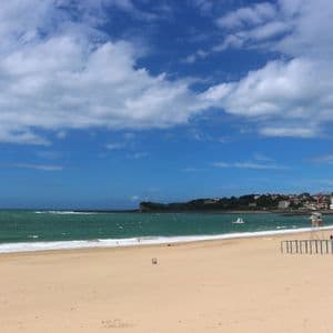 Una spiaggia ampia e sabbiosa incontra il mare verde sotto un cielo blu con nuvole bianche, con una cittadina costiera in lontananza.