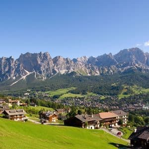 Un villaggio alpino con edifici in legno è immerso in una valle verde ai piedi di una catena montuosa rocciosa.