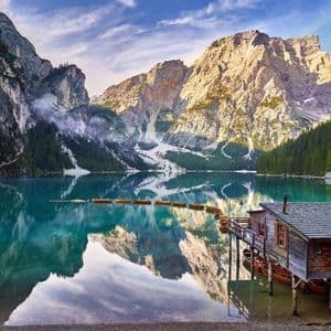 Una caseta de madera y botes de remos en un lago turquesa, con grandes montañas reflejándose en las tranquilas aguas bajo un cielo azul.