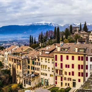 A hillside town with terracotta-roofed buildings nestled below a large, snow-capped mountain range under a cloudy sky.