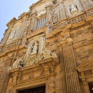 Una ripresa dal basso della facciata in pietra riccamente decorata di una chiesa barocca, con intagli complessi, colonne e statue, sotto un cielo limpido.