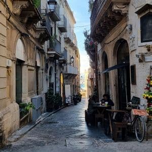 Vista su uno stretto vicolo acciottolato con persone in un caffè all'aperto, che porta al mare in lontananza.