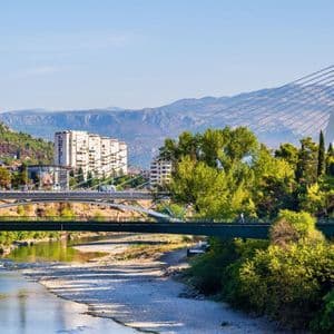 Un moderno ponte strallato bianco e un ponte pedonale verde attraversano un fiume poco profondo che scorre attraverso una città con montagne sullo sfondo.
