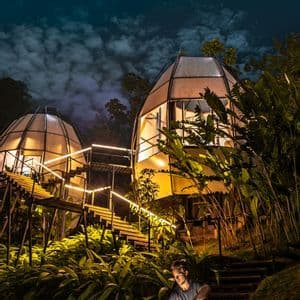 Un uomo siede su una terrazza di legno di notte, con una luce in mano davanti a due cabine a forma di cupola illuminate in una giungla lussureggiante.