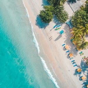 Vista aérea cenital de una playa tropical con agua turquesa, arena blanca, palmeras, kayaks de colores y un edificio con tejado rojo.