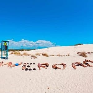 Un viaggio di gruppo WeRoad, sdraiati su una spiaggia di sabbia bianca, che compongono la parola WEROAD con i loro corpi e le borse, sotto un cielo blu.