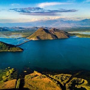 Una veduta dall'alto di un immenso lago blu, circondato da verdi terre, con un ponte che collega una penisola e le montagne in lontananza.