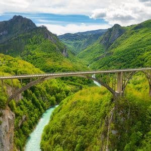Un ponte ad arco attraversa un fiume turchese in una profonda valle, circondata da lussureggianti montagne verdi, sotto un cielo parzialmente nuvoloso.