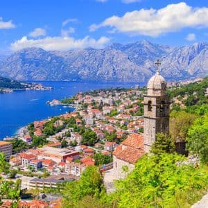 Un campanile in pietra domina una città costiera incastonata tra una baia azzurra e imponenti montagne sotto un cielo soleggiato.