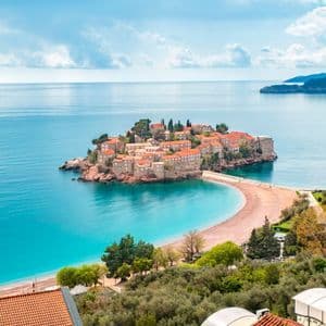 Un isolotto fortificato con tetti in tegole rosse si erge in acque turchesi, collegato a una spiaggia sabbiosa da uno stretto istmo.