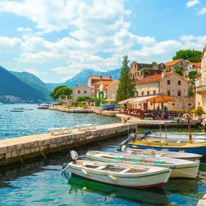 Piccole imbarcazioni sono ormeggiate lungo un molo di pietra in una cittadina di mare, con verdi montagne che si ergono sullo sfondo sotto un cielo blu, parzialmente nuvoloso.