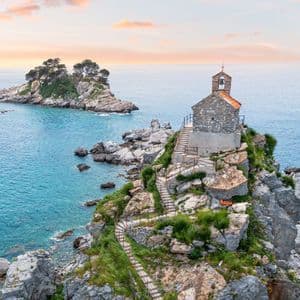 Una vecchia chiesa in pietra con un campanile si erge su un isolotto roccioso nel mare, vista al tramonto.