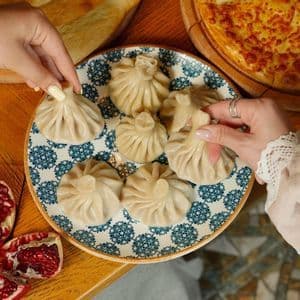Due mani prendono i khinkali da un piatto a fantasia blu e bianca su un tavolo di legno circondato da altri piatti georgiani.