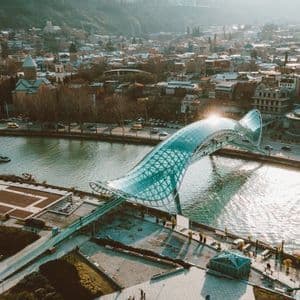 Una veduta aerea di un moderno ponte pedonale in vetro e acciaio che si estende su un fiume, con uno skyline cittadino sullo sfondo.