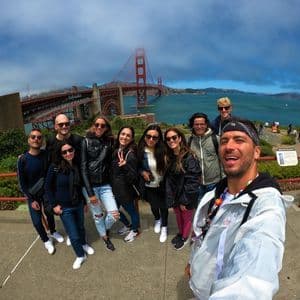 A WeRoad group trip takes a selfie from a viewpoint, with a large red suspension bridge and the sea in the background.