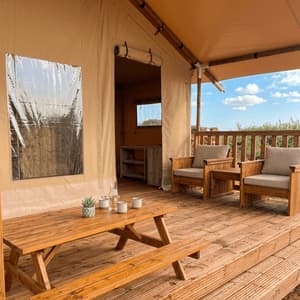 La pedana in legno di una tenda glamping in tela, arredata con un tavolo da picnic e due poltrone sotto una tenda da sole.
