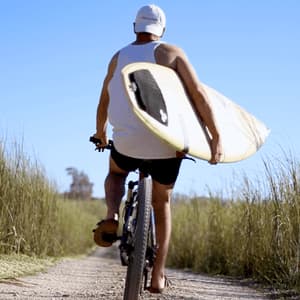 Un uomo visto di spalle pedala in bicicletta su un sentiero sterrato, con una tavola da surf sotto il braccio.