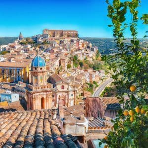 Vista soleggiata dall'alto di una città storica su una collina con una cupola blu, tra tetti in terracotta e un albero di limoni.