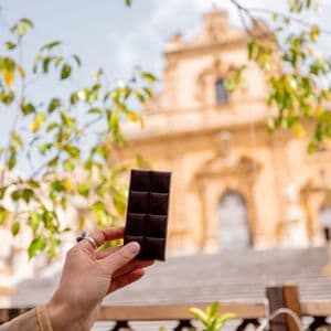 Una mano regge una tavoletta di cioccolato fondente in primo piano, con un edificio ornato e color sabbia sfocato sullo sfondo.