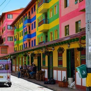 Una strada acciottolata, fiancheggiata da edifici a strisce dai colori vivaci, con un veicolo viola parcheggiato e persone sedute ai tavolini all'aperto.