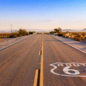 The Route 66 shield painted on a long, empty asphalt road stretching through a desert landscape at sunset.