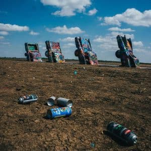 Graffiti-covered cars buried nose-down in a field, with empty spray paint cans on the cracked earth under a blue, cloudy sky.