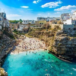 Una vista dall'alto di una spiaggia affollata in una caletta con acqua turchese, circondata da scogliere sormontate da edifici.